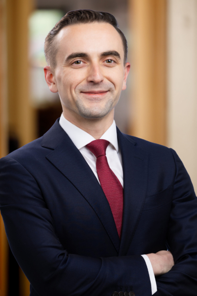 Professional headshot of a smiling man in a navy suit and red tie with arms crossed in a corporate setting.